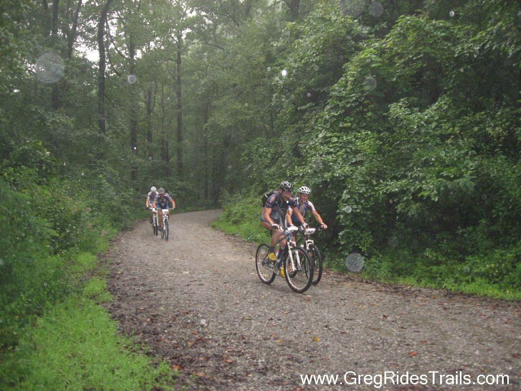 Three mountain bikers ride along a gravel trail in a lush, green forest on a rainy day. The path curves gently ahead, surrounded by dense foliage and trees. Raindrops can be seen in the air, adding to the damp atmosphere. Winding Stairs Loop mountain bike trail.