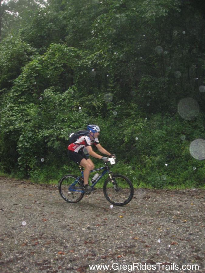 A cyclist riding a mountain bike on a gravel path during a rainstorm, with rain droplets visible in the air and a dense, green forest in the background. The rider is wearing a helmet and a biking outfit, indicating an active outdoor setting. Winding Stairs Loop mountain bike trail.