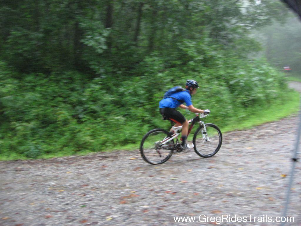 A mountain biker in a blue shirt and black shorts is riding swiftly on a gravel trail surrounded by dense greenery, with misty conditions and a blurred background indicating speed. Winding Stairs Loop mountain bike trail.