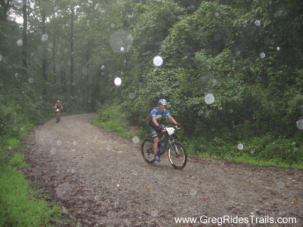 Two cyclists navigating a gravel path in a dense forest during rain, with droplets visible in the air. The first cyclist is wearing an orange shirt, while the second is in a blue jersey, focusing on the trail ahead amidst a misty, wet environment. Winding Stairs Loop mountain bike trail.