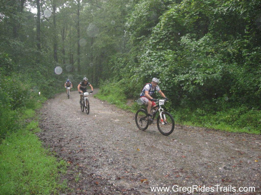 Three mountain bikers compete on a gravel trail in a forest during rain, with trees and greenery surrounding the path. Raindrops are visible in the air, adding to the atmosphere of the challenging ride. Winding Stairs Loop mountain bike trail.