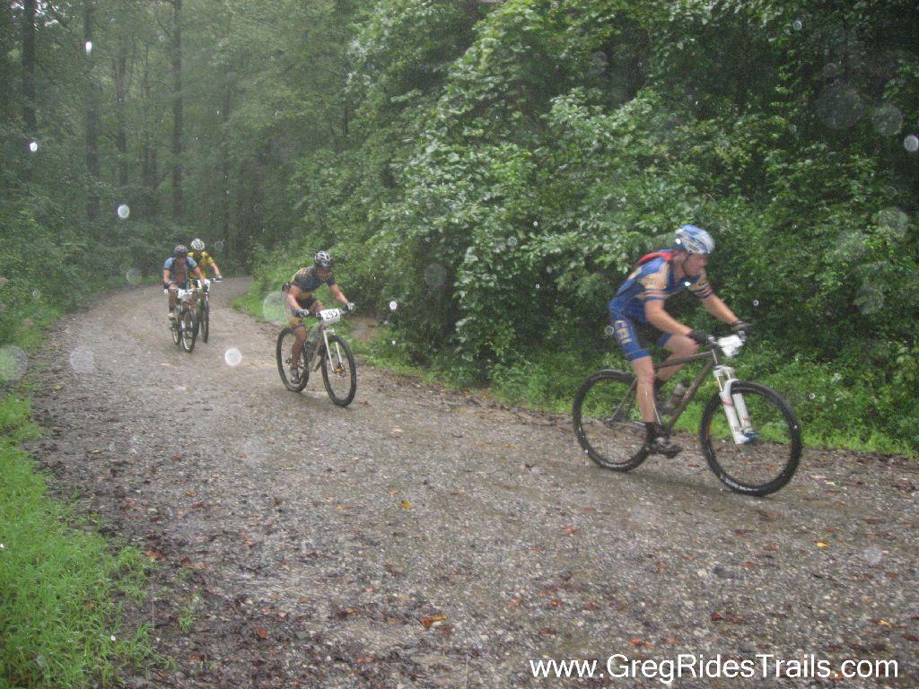 Four mountain bikers navigate a muddy, gravel path in a lush, green forest during rainy weather. The riders, dressed in colorful cycling gear, appear focused as they maneuver through the challenging conditions. Rain can be seen in the foreground, adding to the dynamic atmosphere of the scene. Winding Stairs Loop mountain bike trail.
