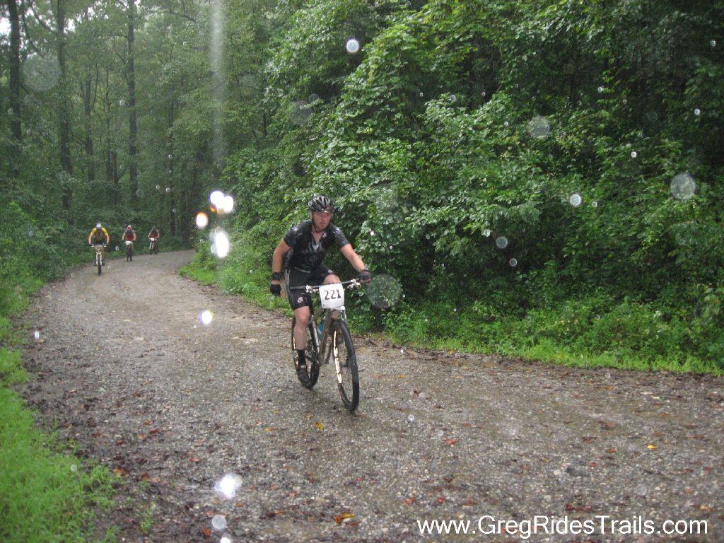 Mountain bikers ride along a gravel trail in a lush, green forest during a rainstorm. One rider in the foreground, wearing a black jersey and helmet, is focused on the path ahead while others trail behind. The scene captures the challenging conditions of outdoor cycling in wet weather. Winding Stairs Loop mountain bike trail.