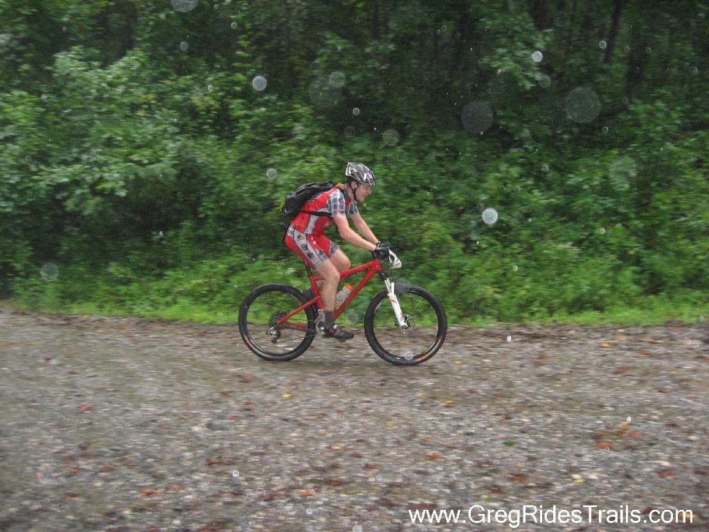 A mountain biker in a red cycling outfit rides on a gravel trail during rainy weather, with greenery in the background and rain droplets visible in the air. Winding Stairs Loop mountain bike trail.