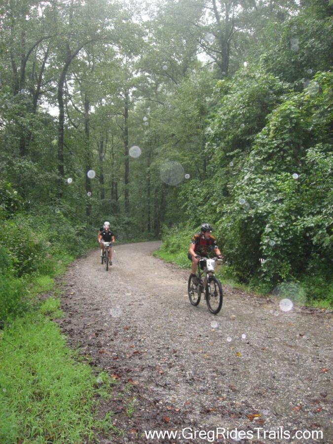 Two mountain bikers ride along a gravel path surrounded by lush greenery in a rainy forest setting. Raindrops are visible in the air, adding to the wet atmosphere. Winding Stairs Loop mountain bike trail.