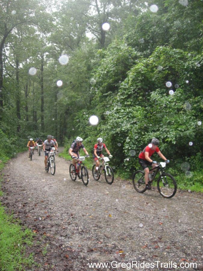 A group of mountain bikers riding on a gravel trail through a lush green forest, with rain falling heavily and creating splashes. The cyclists are wearing helmets and gear, demonstrating determination as they navigate the slippery terrain. Large droplets of rain can be seen in the foreground, adding to the atmosphere of the scene. Winding Stairs Loop mountain bike trail.