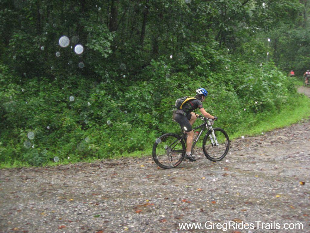 A mountain biker rides along a gravel trail in the rain, with large raindrops visible in the air and lush green foliage lining the path. Winding Stairs Loop mountain bike trail.