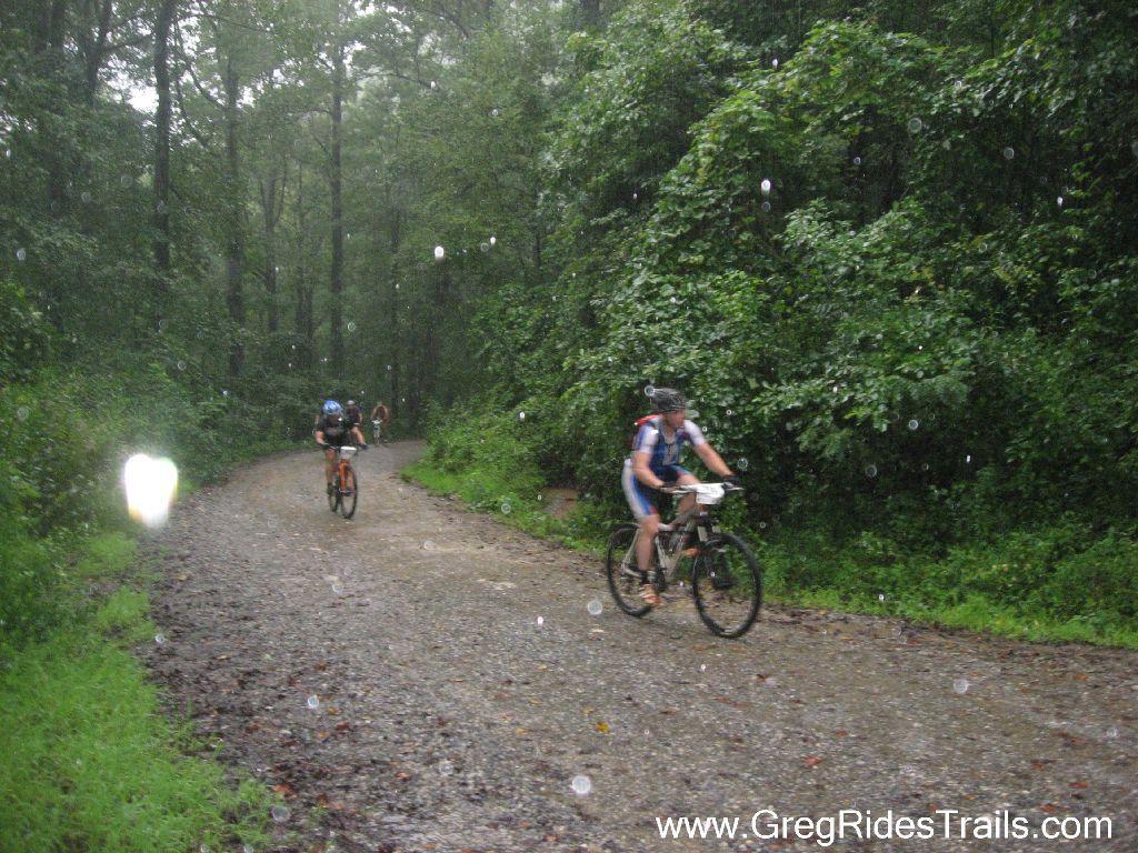 Two mountain bikers navigate a gravel trail in a forest during rain, with droplets visible in the air. The surrounding greenery is vibrant and lush, enhancing the outdoor atmosphere. Winding Stairs Loop mountain bike trail.