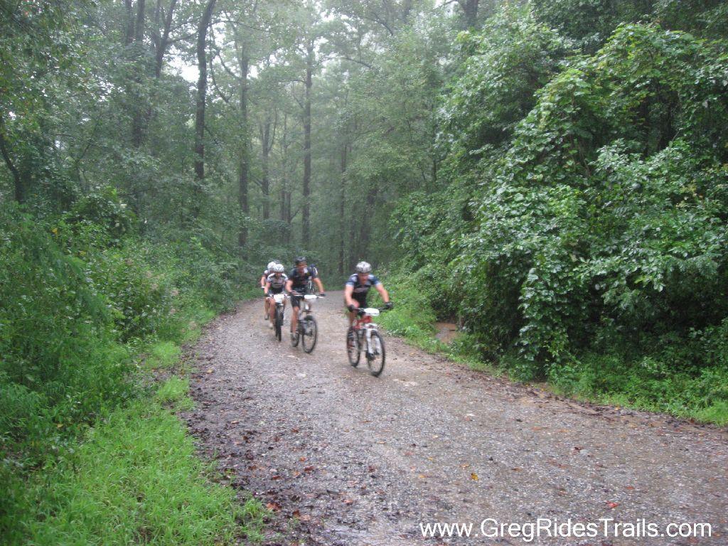 Four mountain bikers riding on a wet, gravel trail surrounded by dense greenery and trees during rainy weather. Winding Stairs Loop mountain bike trail.