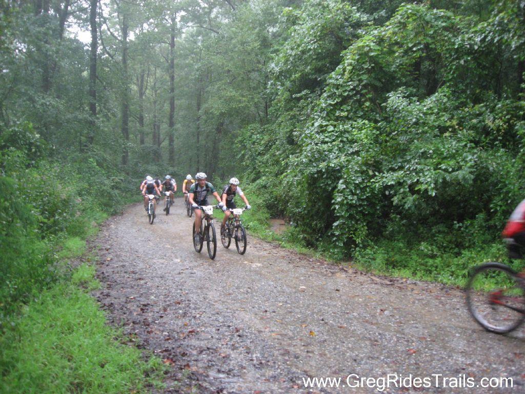 Mountain bikers riding on a gravel trail surrounded by lush green forest during rainy weather. The path is slightly muddy, and several cyclists are visible, wearing helmets and cycling gear, as they navigate the terrain. Winding Stairs Loop mountain bike trail.