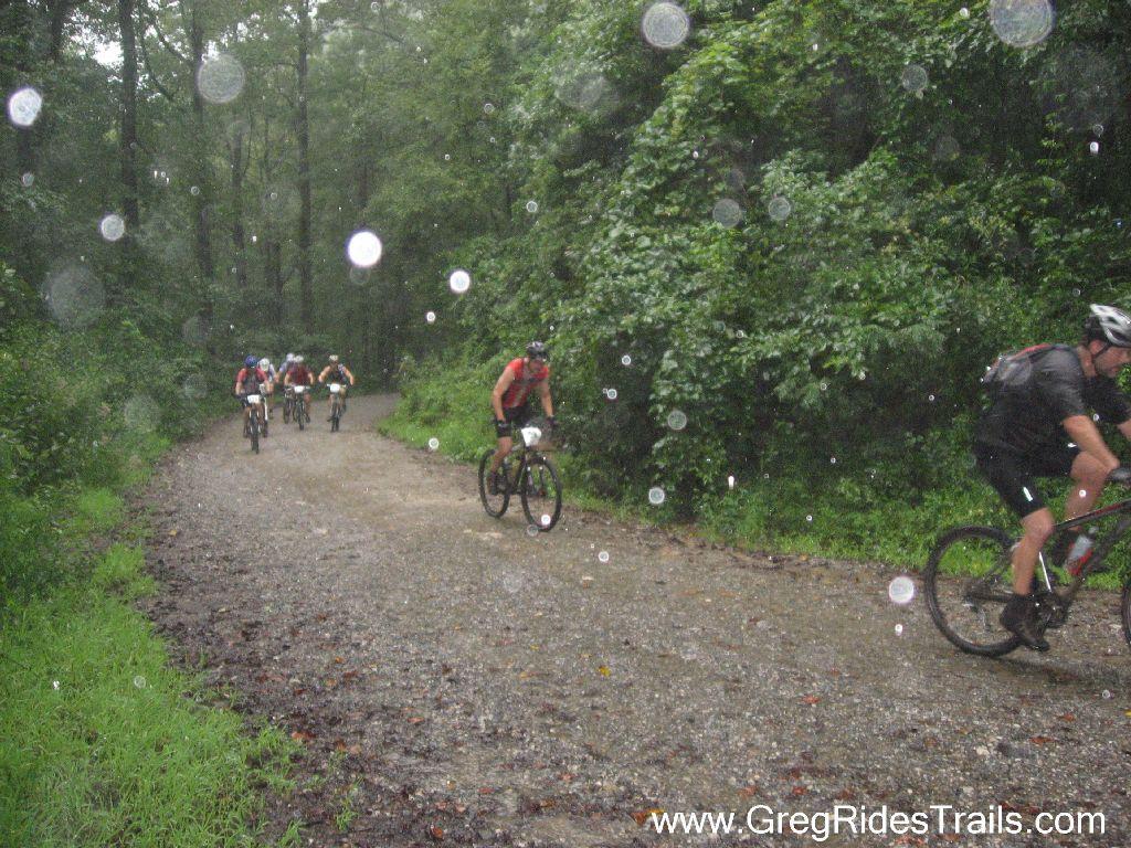 A group of mountain bikers traverses a muddy, gravel trail surrounded by lush greenery, as rain falls heavily, creating splashes on the ground. The scene captures the challenging conditions of the ride, with several riders visible in the background and one cyclist in the foreground. Winding Stairs Loop mountain bike trail.