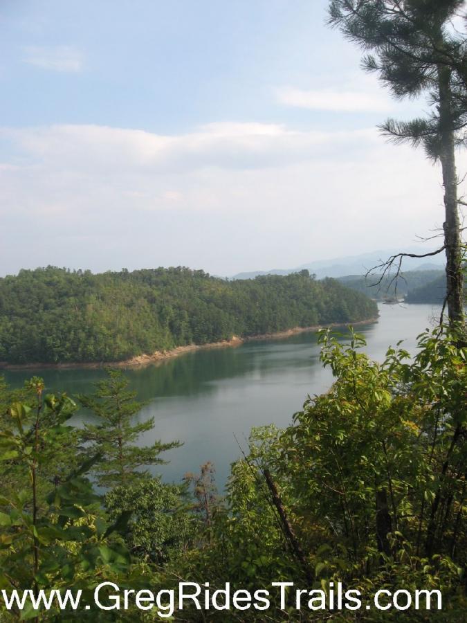 A scenic view of a calm lake surrounded by lush green hills and trees under a partly cloudy sky. The image captures the tranquility of nature, with the water reflecting the landscape. Tsali Right Loop mountain bike trail.