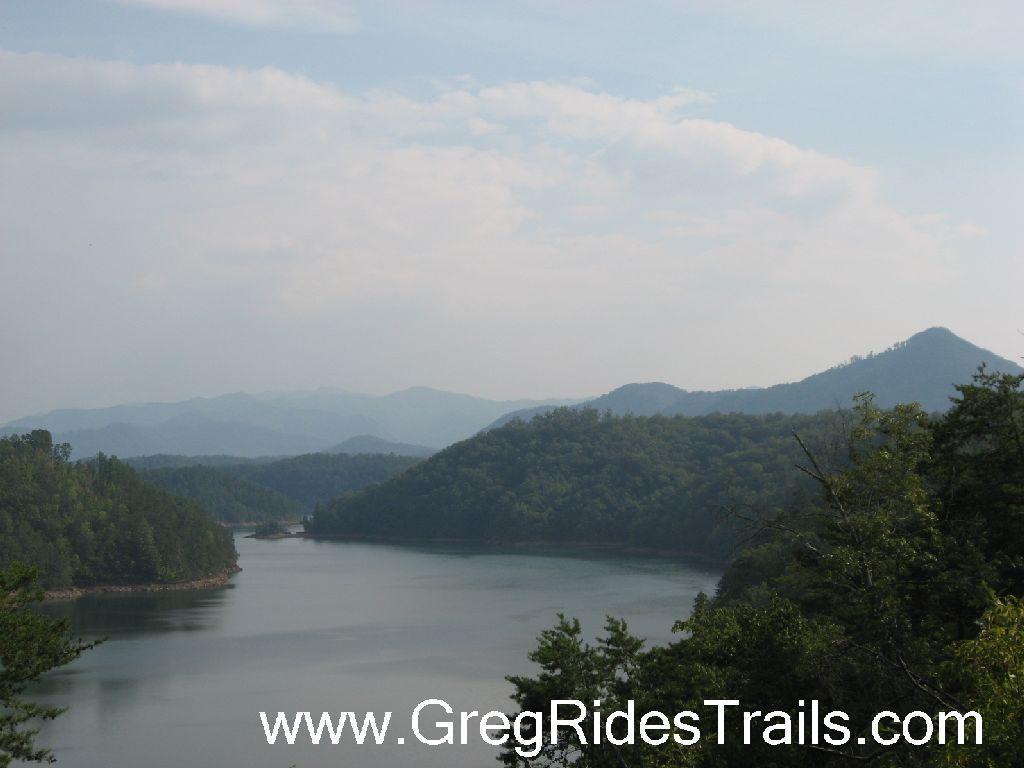 A serene landscape featuring a calm lake surrounded by lush green hills and mountains under a cloudy sky. The water reflects the surrounding nature, creating a peaceful outdoor scene. Tsali Recreation Area mountain bike trail.