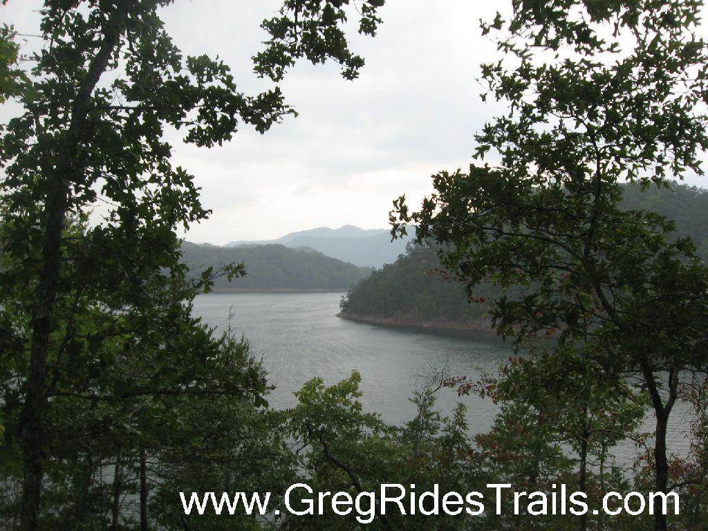 A tranquil view of a serene lake surrounded by dense greenery and mountains in the background. The sky is overcast, enhancing the peaceful atmosphere of the natural landscape. Tsali Recreation Area mountain bike trail.