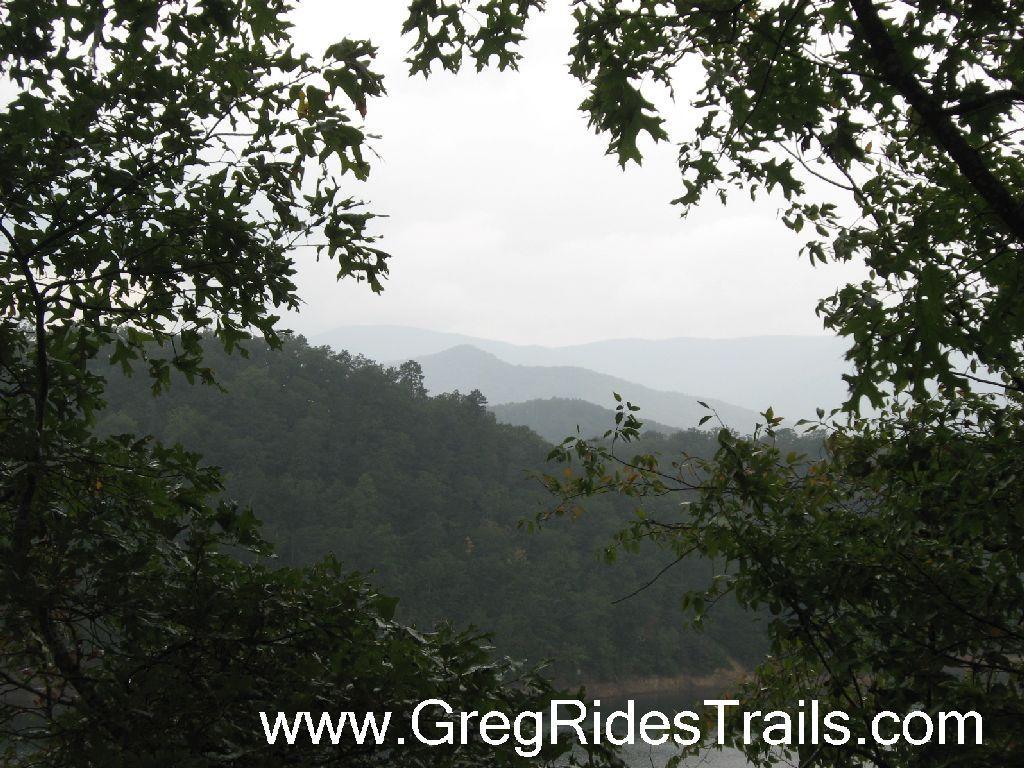 A scenic view of rolling green hills and distant mountains framed by lush foliage under a cloudy sky. Tsali Recreation Area mountain bike trail.