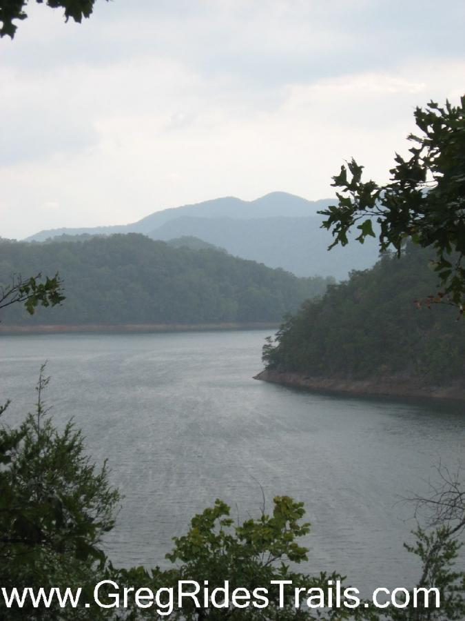 A serene view of a winding lake surrounded by lush green hills and distant mountains under a cloudy sky. The water appears calm, reflecting the natural landscape. Tsali Left Loop mountain bike trail.