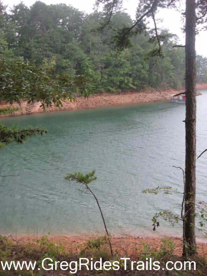 A serene view of a calm lake surrounded by lush green trees. The water reflects a tranquil blue hue, with a sandy shore visible in the background. A small boat is docked at the water's edge, adding to the peaceful atmosphere of this natural setting. Tsali Recreation Area mountain bike trail.