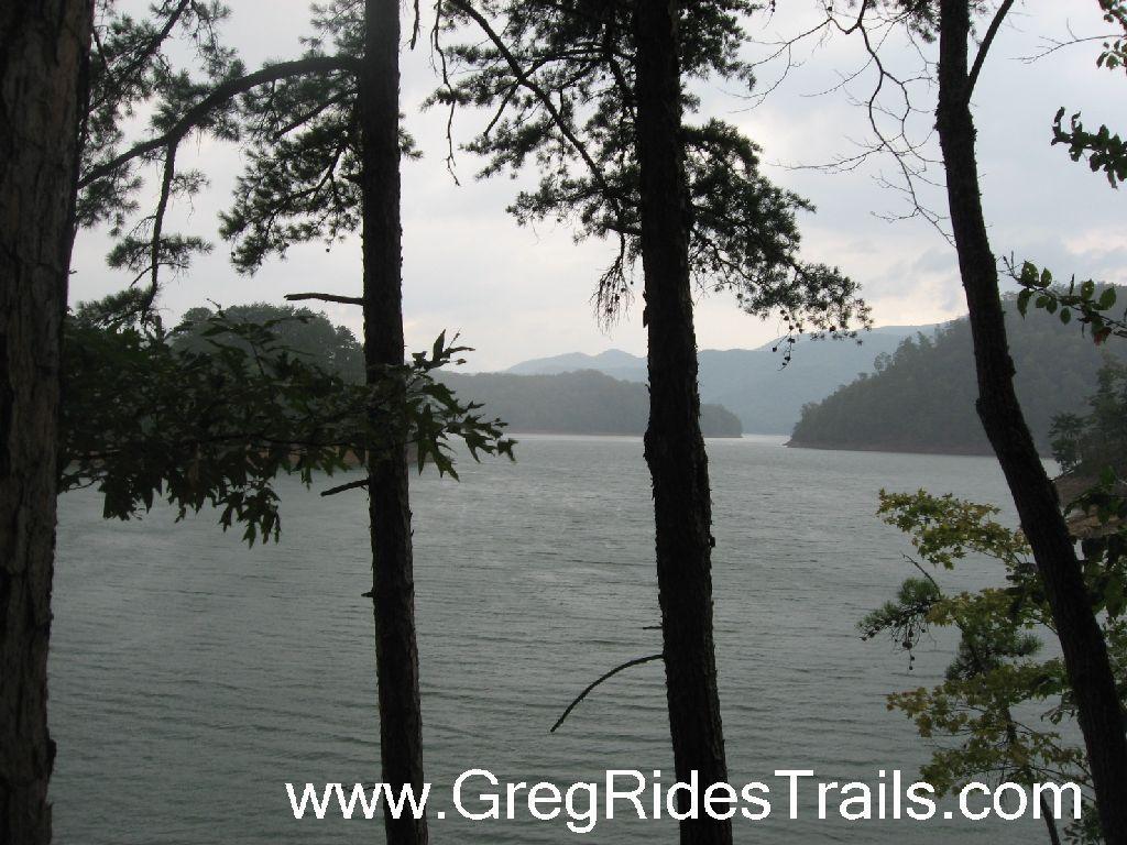A serene landscape view of a calm lake surrounded by trees, with distant mountains under a cloudy sky. The image captures a tranquil outdoor setting, perfect for nature exploration. Tsali Recreation Area mountain bike trail.