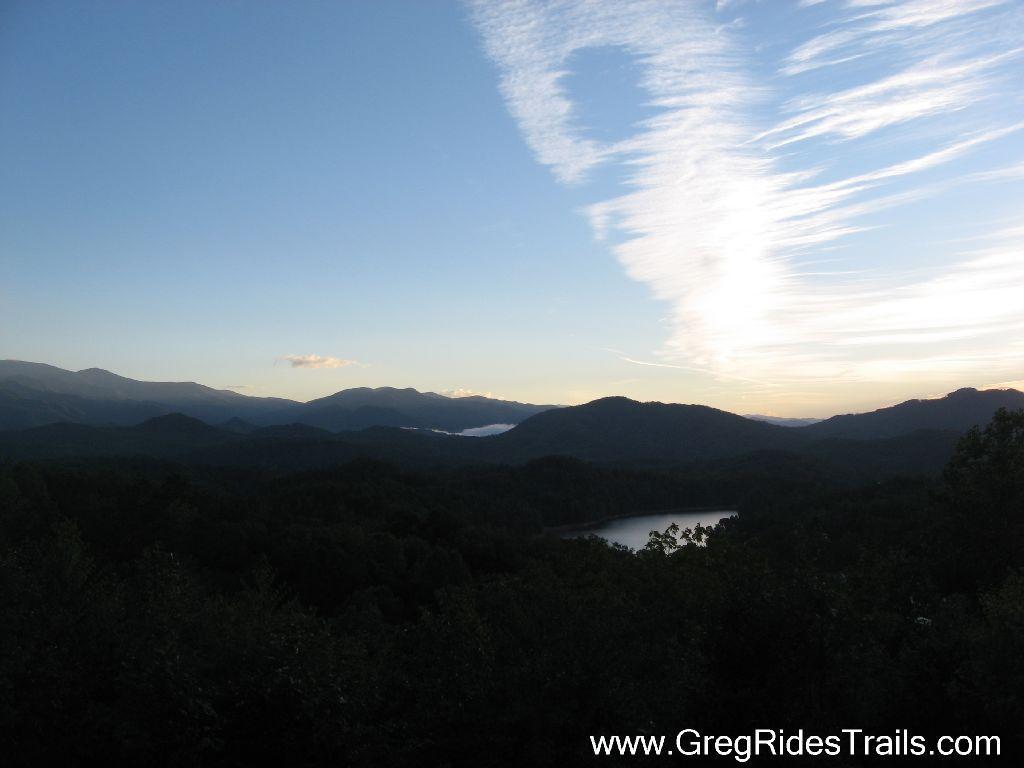 A scenic view of a mountainous landscape during twilight, featuring layered mountains in the background and a tranquil lake nestled among trees in the foreground. The sky is partially covered with wispy clouds, transitioning from daylight to evening. Tsali Recreation Area mountain bike trail.