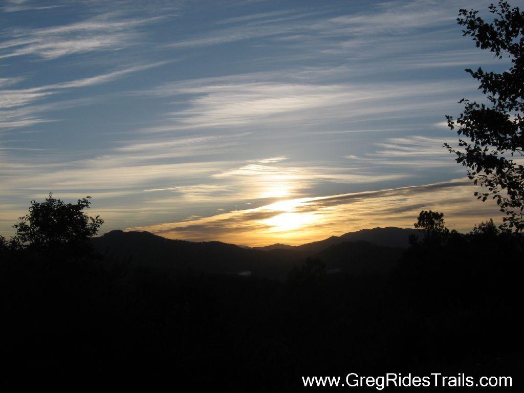 Sunset over a mountain landscape, with a colorful sky featuring wispy clouds and warm hues reflecting the setting sun. Silhouettes of trees and hills create a serene natural scene. Tsali Recreation Area mountain bike trail.