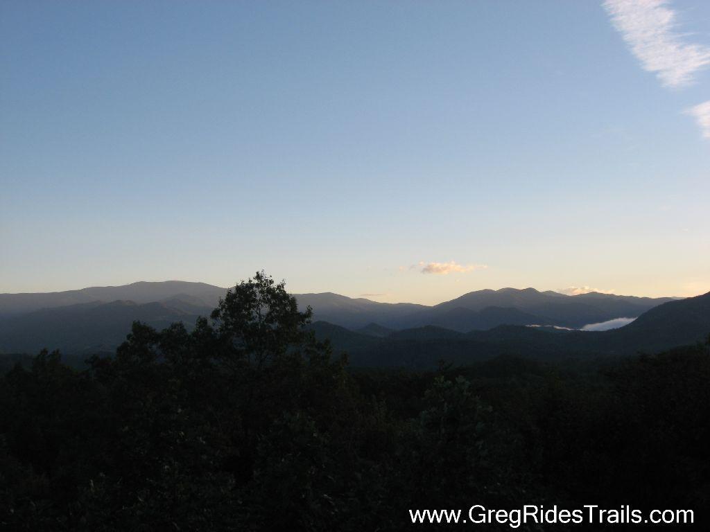 A scenic view of mountain ranges under a clear sky at dusk, with lush greenery in the foreground and soft clouds lingering in the valleys. Tsali Recreation Area mountain bike trail.