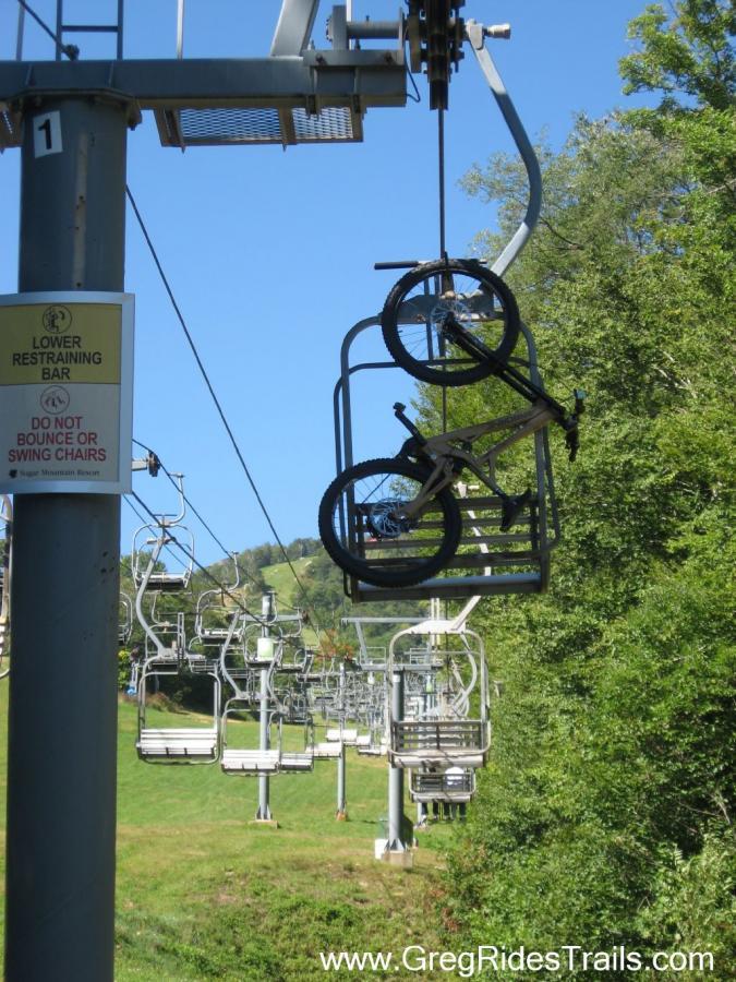 A bike hanging on a ski lift chair with a clear blue sky in the background, surrounded by green trees and a ski slope visible in the distance. A sign nearby warns not to bounce or swing chairs. Sugar Mountain Resort mountain bike trail.