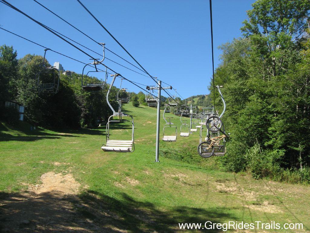 A scenic view of a ski lift on a clear day, with empty chairs ascending a grassy hill surrounded by trees. The sky is bright blue, indicating a sunny weather. Some bike carriers are visible on a few lift chairs, suggesting options for mountain biking. Sugar Mountain Resort mountain bike trail.