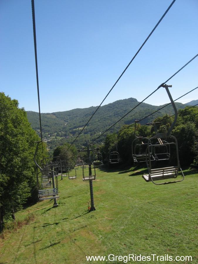 A scenic view from a ski lift, showing empty ski chairs hanging on cables above a green grassy slope. In the background, rolling hills are visible under a clear blue sky. Sugar Mountain Resort mountain bike trail.