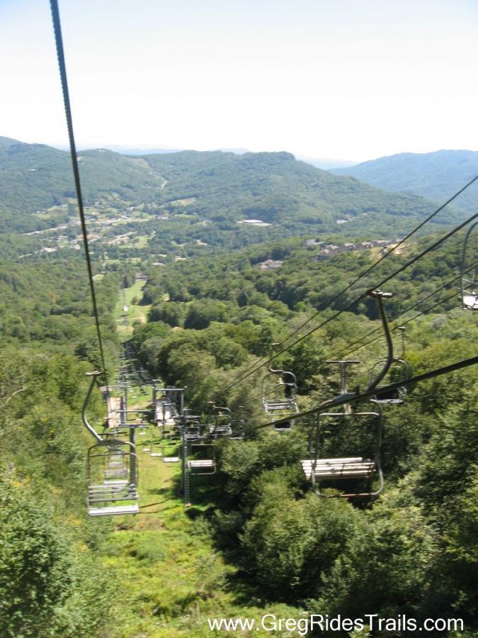 A scenic view from a chairlift overlooking a green valley surrounded by mountains, with ski trails visible below and a clear blue sky above. Sugar Mountain Resort mountain bike trail.