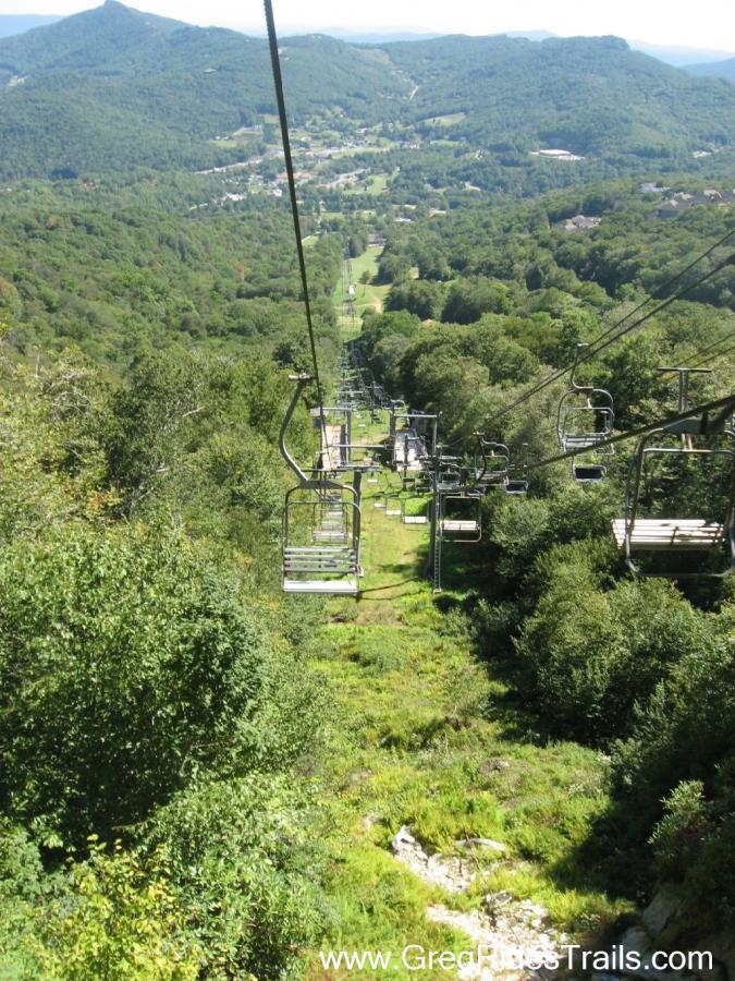 A scenic view from a ski lift, overlooking a lush, green valley surrounded by mountains. The image captures the chairlift cables and empty seats descending through dense trees, with a winding path visible below leading into the valley. Sugar Mountain Resort mountain bike trail.