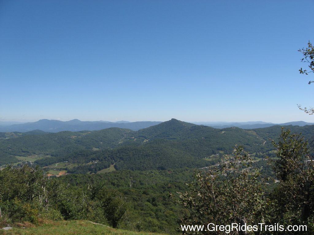 A panoramic view of lush green mountain ranges under a clear blue sky, showcasing rolling hills and valleys in the foreground. The landscape features a mix of wooded areas and open fields, with distant mountains fading into the horizon. Sugar Mountain Resort mountain bike trail.