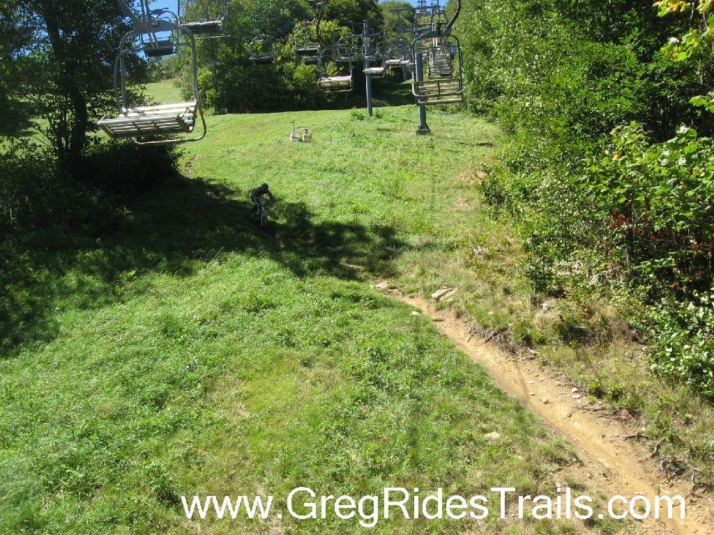 A scenic view of a grassy hillside with a mountain bike trail winding down. In the background, a ski lift with empty chairs is visible, surrounded by lush green trees and a clear blue sky. The area suggests a recreational site for biking or skiing. Sugar Mountain Resort mountain bike trail.