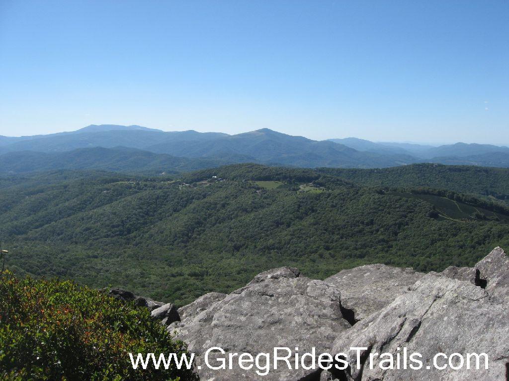 A panoramic view of rolling green mountains under a clear blue sky, taken from a rocky viewpoint. The landscape features layered hills and valleys, with patches of forest and farmland visible in the distance. Sugar Mountain Resort mountain bike trail.