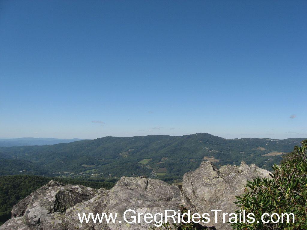 A panoramic view of lush green mountains under a clear blue sky, with rocky outcrops in the foreground. The landscape features rolling hills and valleys, showcasing the natural beauty of the area. Sugar Mountain Resort mountain bike trail.