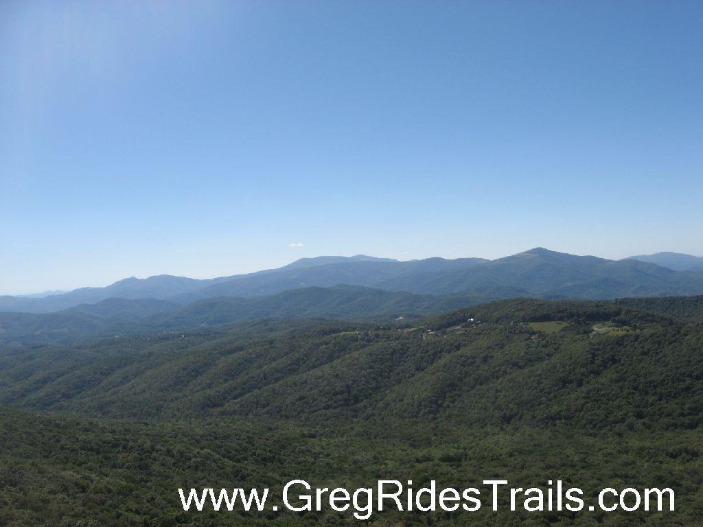 A panoramic view of rolling green hills and distant mountains under a clear blue sky. The landscape features layers of mountains fading into the horizon, showcasing a natural, serene environment. Sugar Mountain Resort mountain bike trail.