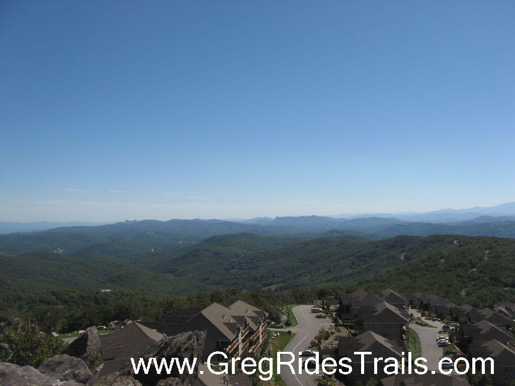 A panoramic view of rolling green hills and distant mountains under a clear blue sky, featuring residential houses in the foreground. The landscape showcases a mix of trees and winding roads, highlighting the natural beauty of the area. Sugar Mountain Resort mountain bike trail.