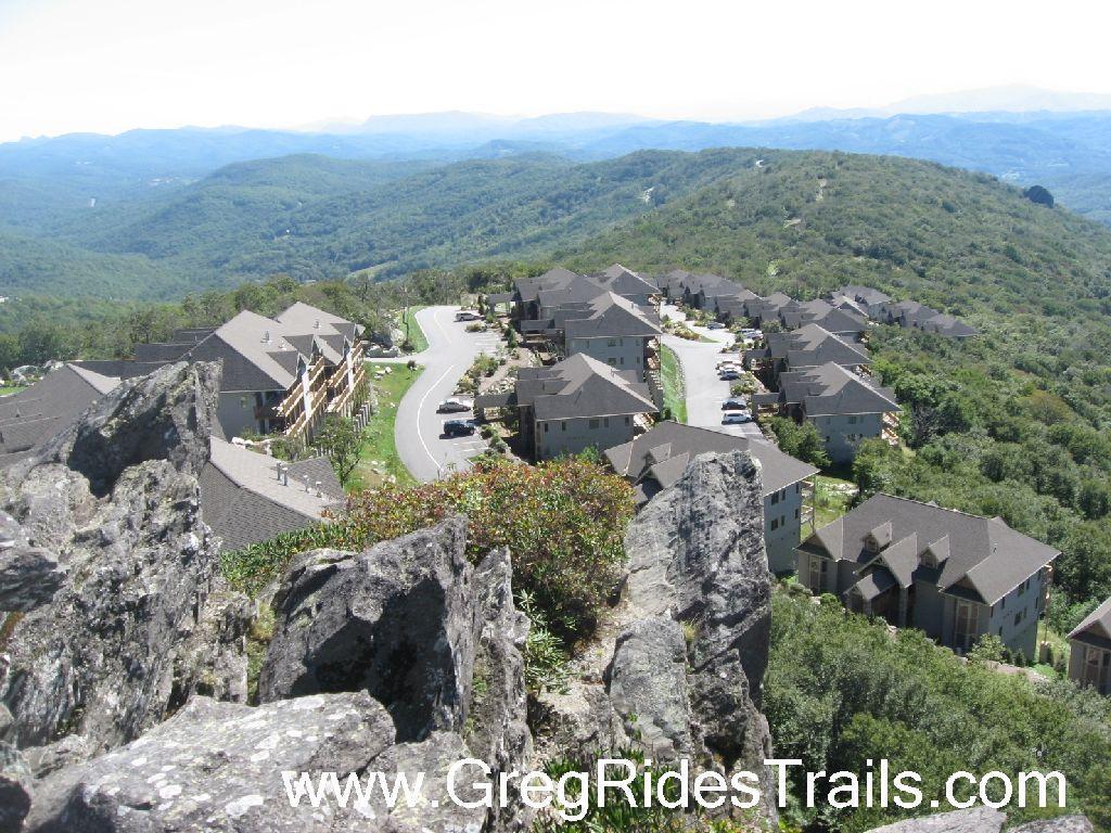 Aerial view of a scenic mountain landscape featuring rocky outcrops in the foreground, with a residential area and winding road visible below. Lush green hills extend into the distance under a clear sky. Sugar Mountain Resort mountain bike trail.