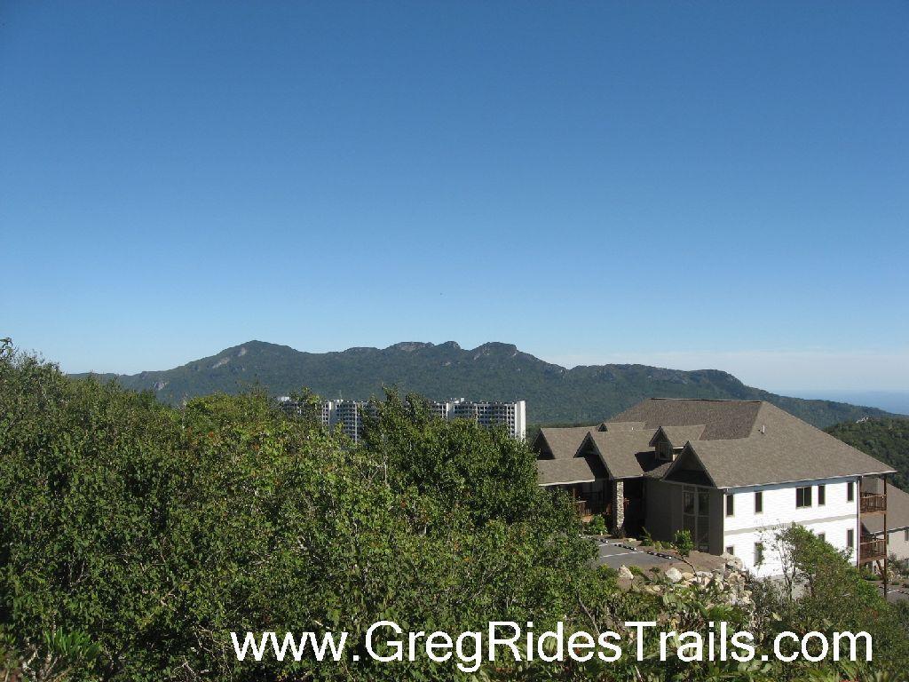 A scenic view of mountains under a clear blue sky, with a foreground featuring lush greenery and a building with a sloped roof. Sugar Mountain Resort mountain bike trail.