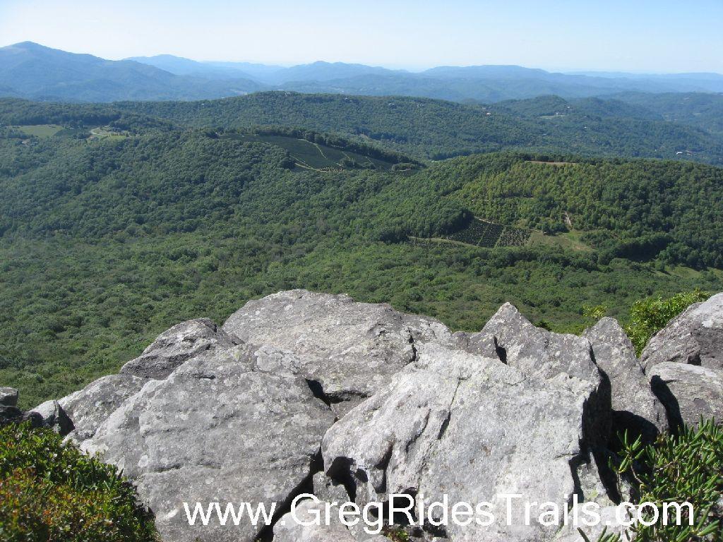 A panoramic view of rolling green mountains and valleys, taken from a rocky overlook. The foreground features rugged grey rocks, while the background showcases lush forests interspersed with patches of cultivated land. Clear blue skies are visible above, creating a serene and natural landscape. Sugar Mountain Resort mountain bike trail.
