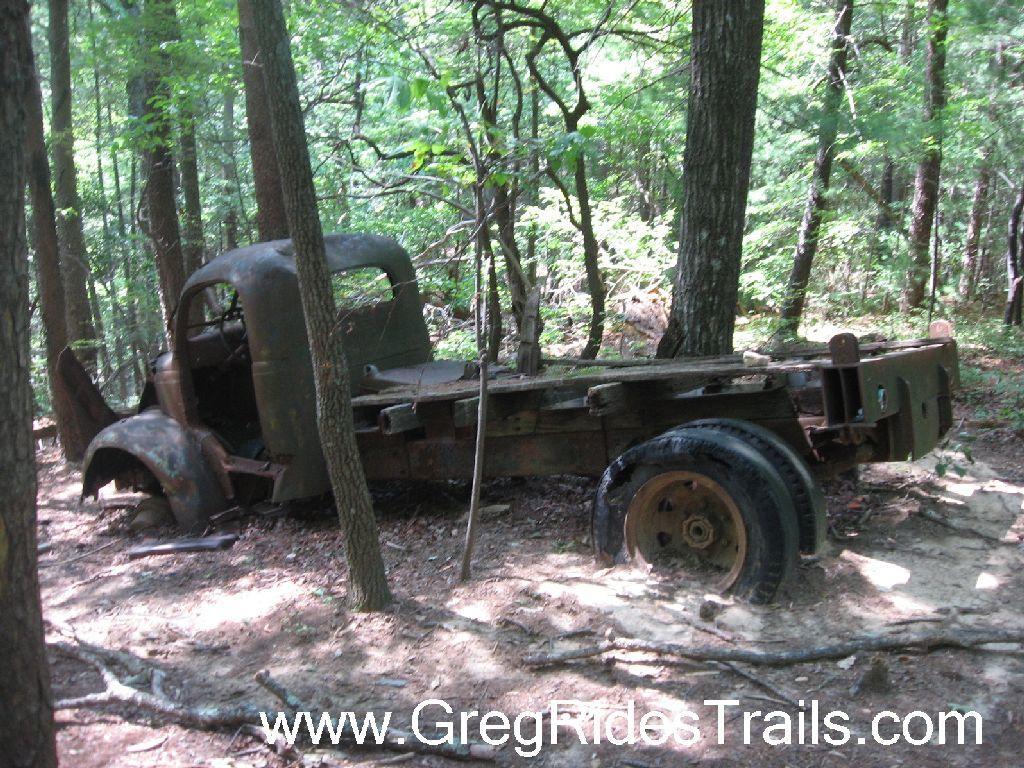 An old, rusty truck abandoned in a wooded area, partially obscured by trees and underbrush. The truck has a weathered exterior with visible signs of aging and decay, showcasing its vintage design. Sunlight filters through the leaves, illuminating the serene natural surroundings. Bull Mountain / 223 mountain bike trail.