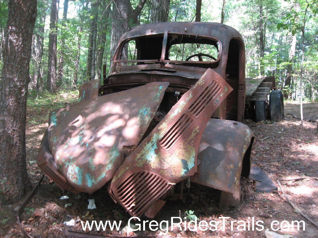 An old, rusted truck partially hidden among trees in a forested area, with peeling paint and a weathered appearance, showcasing the effects of time and nature on vehicles. Bull Mountain / 223 mountain bike trail.