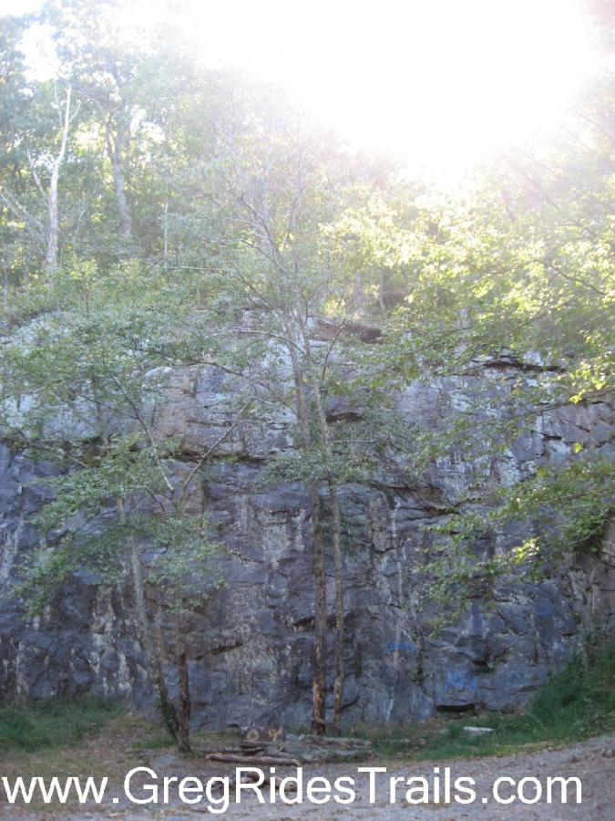 A rocky cliff surrounded by trees, illuminated by bright sunlight filtering through the leaves. The image captures a natural landscape, showcasing the textures of the rock and the greenery of the surrounding foliage. Winding Stairs Loop mountain bike trail.