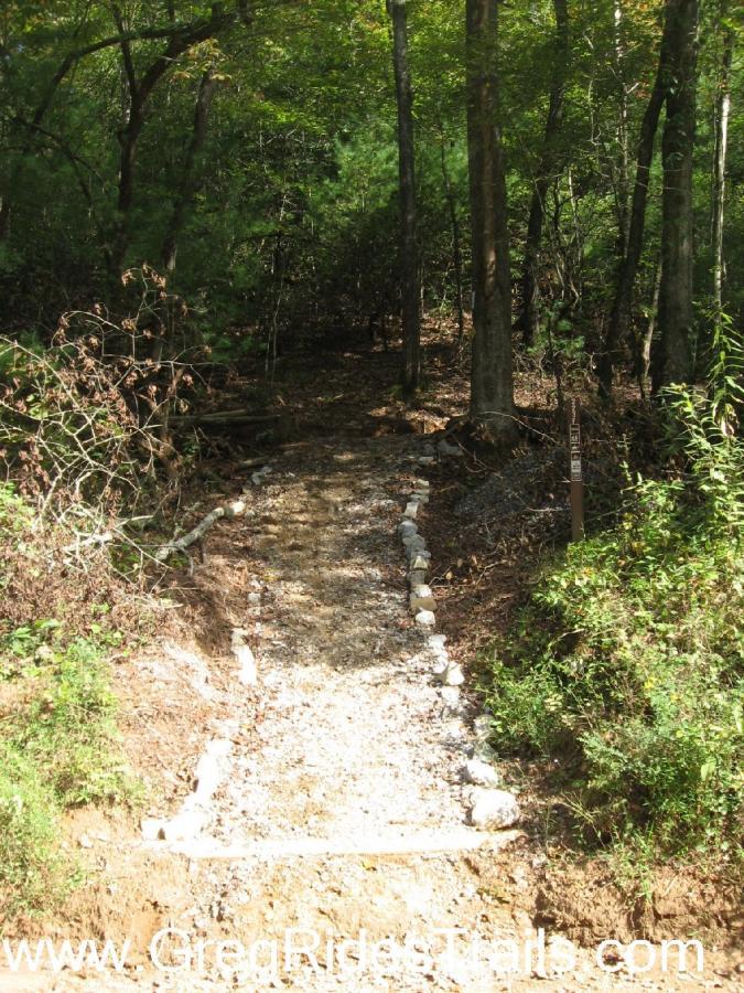 A gravel path leads into a wooded area, bordered by trees and overgrown foliage. The path is lined with small stones, guiding visitors into the natural surroundings. A trail marker is visible on the right side of the image. Jake to Bull Mountain Connecter mountain bike trail.