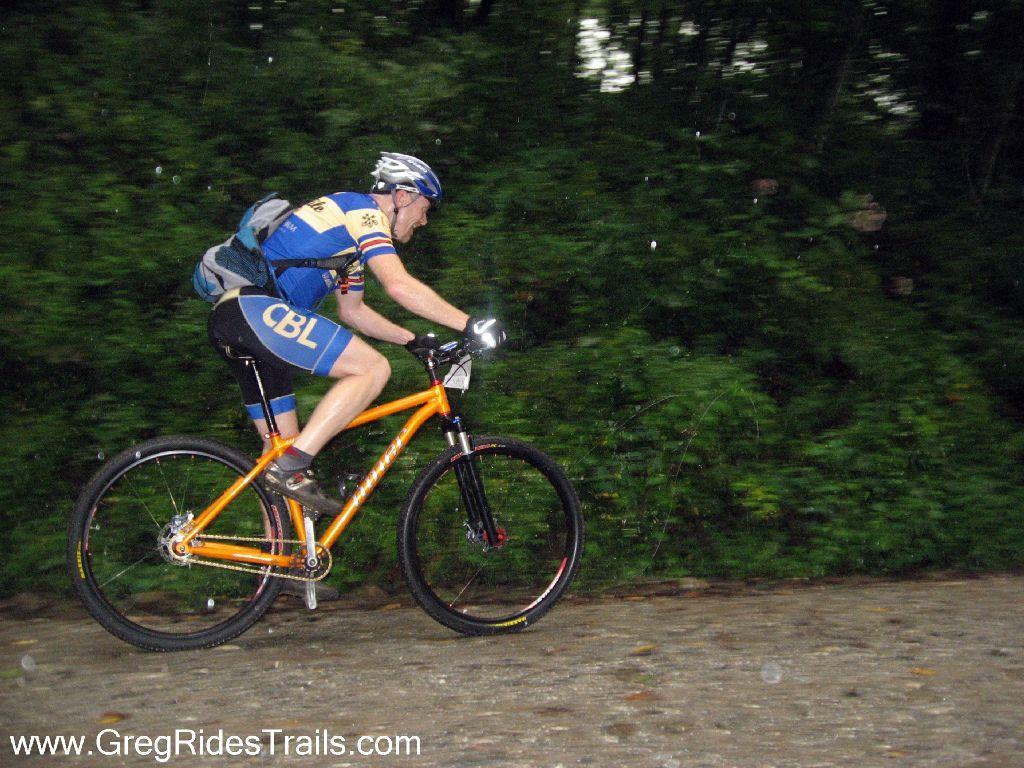 A mountain biker in a blue and yellow jersey rides an orange bike along a gravel path, surrounded by lush green foliage. The rider is focused and appears to be navigating the terrain carefully, with light rain visible in the image. Winding Stairs Loop mountain bike trail.