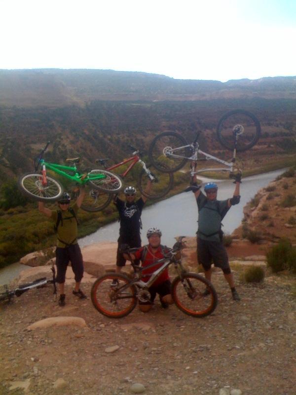Four mountain bikers celebrating at the edge of a canyon, raising their bicycles in triumph. The scenic background features a winding river and rugged terrain under a partly cloudy sky. Two bikes are held above their heads, while one biker kneels beside his bike, all showing enthusiasm for their outdoor adventure. Mary's Loop / Horsethief Bench mountain bike trail.