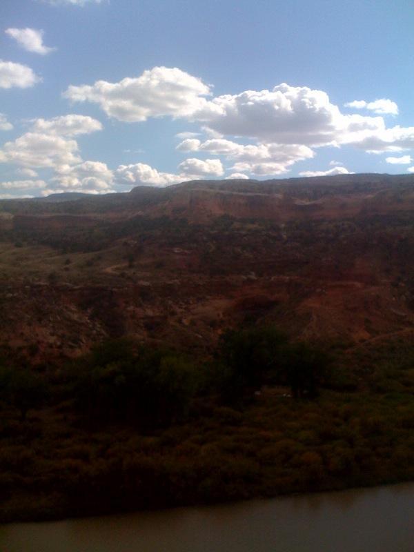 A picturesque landscape featuring rugged cliffs and rolling hills against a blue sky filled with fluffy white clouds. In the foreground, a river winds through dense vegetation, adding to the natural beauty of the scene. Mary's Loop / Horsethief Bench mountain bike trail.