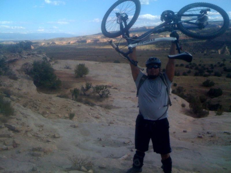 A man wearing a helmet and sunglasses stands triumphantly on uneven terrain, holding a mountain bike above his head. The background features a wide, open landscape with rolling hills and sparse vegetation under a partially cloudy sky. Mary's Loop / Horsethief Bench mountain bike trail.
