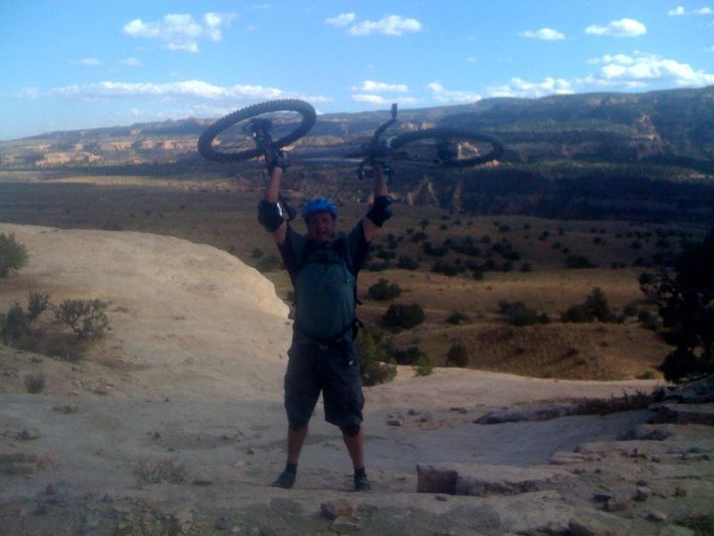 A mountain biker celebrates on a rocky outcrop, holding two mountain bikes above his head, with a vast landscape of hills and valleys in the background under a blue sky with scattered clouds. Mary's Loop / Horsethief Bench mountain bike trail.
