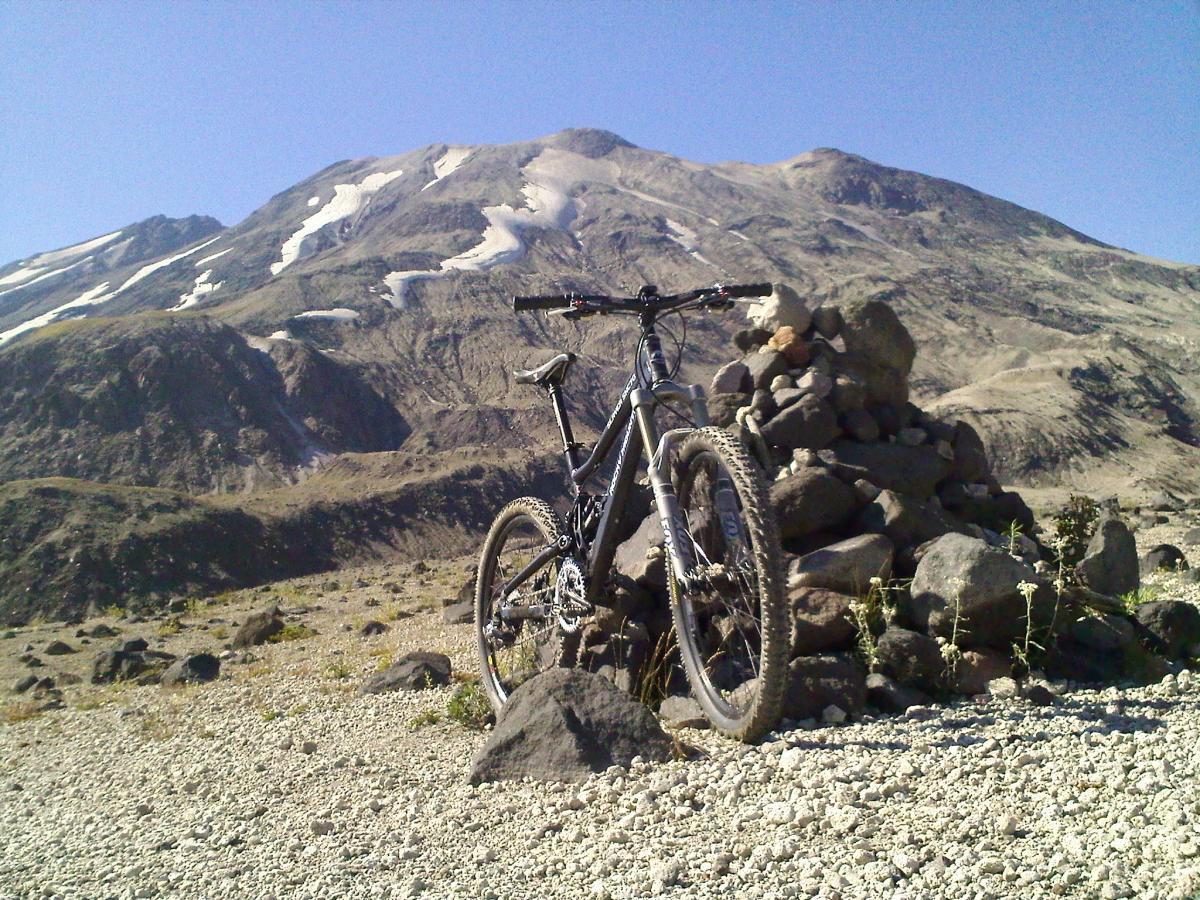 A mountain bike rests on rocky terrain with a mountain range in the background. The foreground features a small stone cairn, while snow-capped peaks rise against a clear blue sky. Ape Canyon#234, Abraham#216d, Smith Creek#225 Trails mountain bike trail.