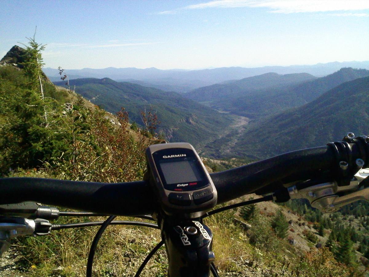 A close-up view of a mountain bike handlebar with a GPS device positioned on it, overlooking a vast mountain valley and ridges in the background under a clear blue sky. Ape Canyon#234, Abraham#216d, Smith Creek#225 Trails mountain bike trail.